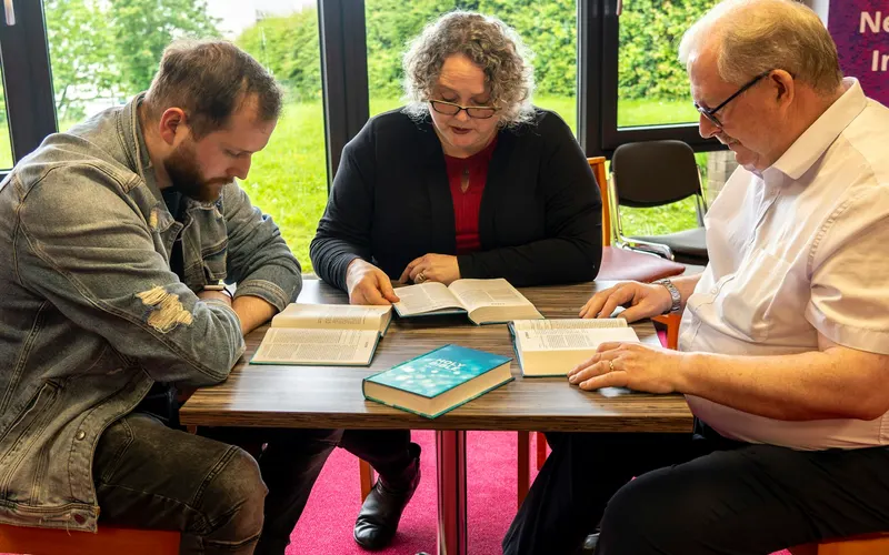 Three adults studying their Bibles together at a table by a window