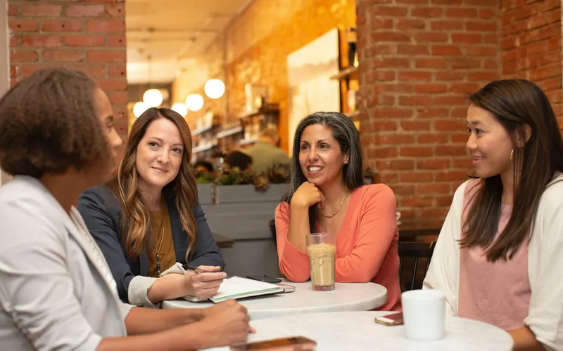 Four women laughing together over coffee at a cafe table