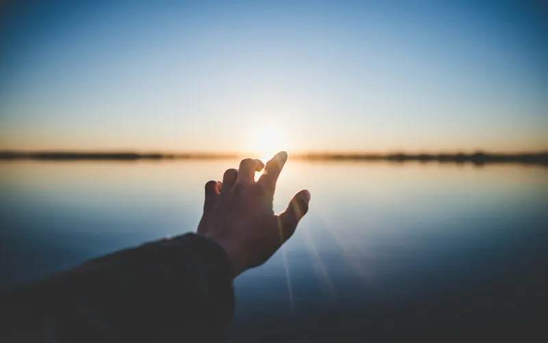 A hand reaching toward the sunrise over still water