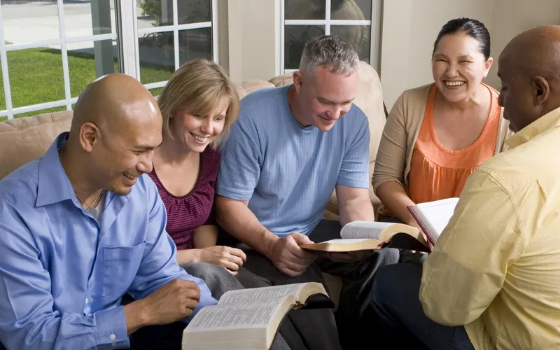 Five friends laughing over open Bibles on the couches of a sunlit living room