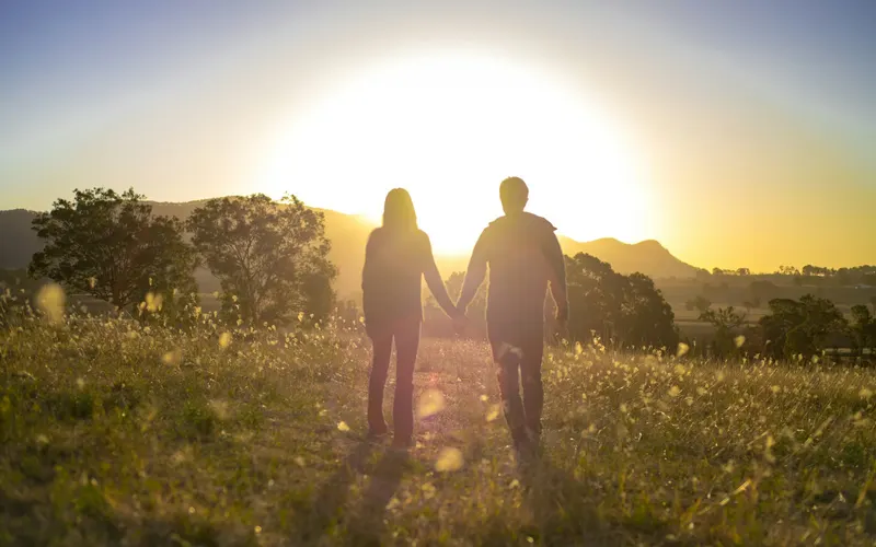 A couple holding hands, walking through a meadow toward the sunset