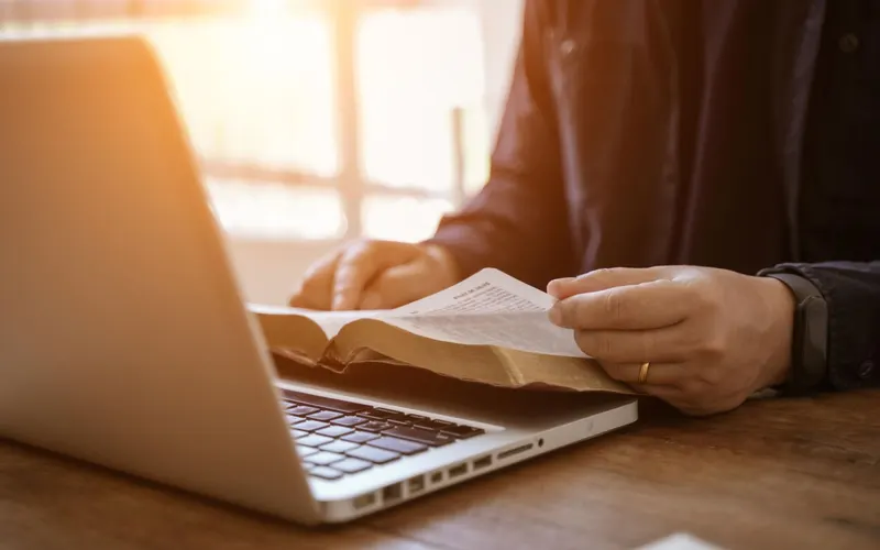 Turning the pages of a Bible beside an open laptop