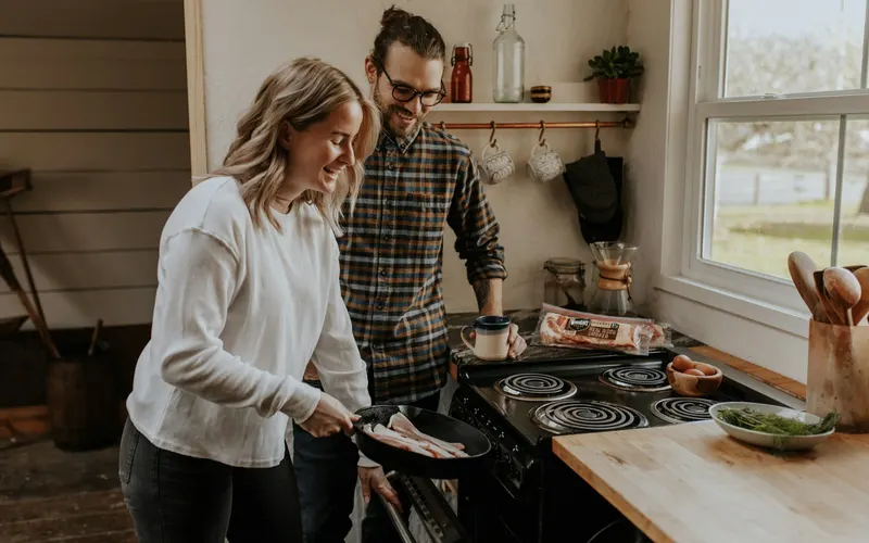 A couple cooking together at a stove by a bright window