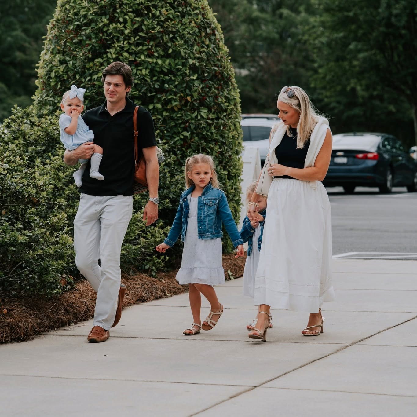 A young family of five walking up the sidewalk to church on a Sunday morning, dad carrying the baby