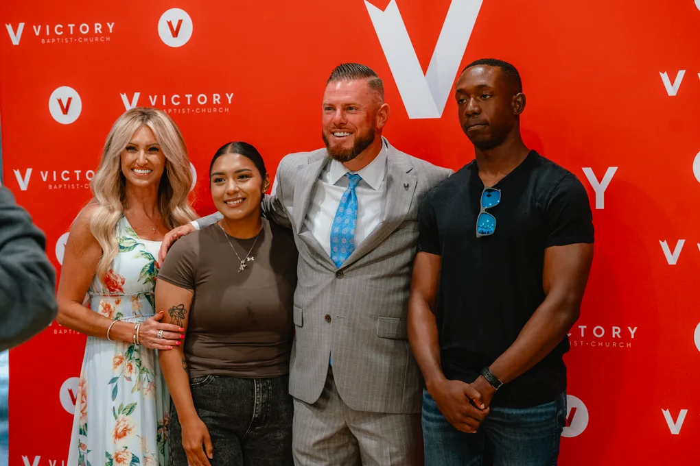 Pastor CT and Becky posing with two guests at the red Victory backdrop after a service