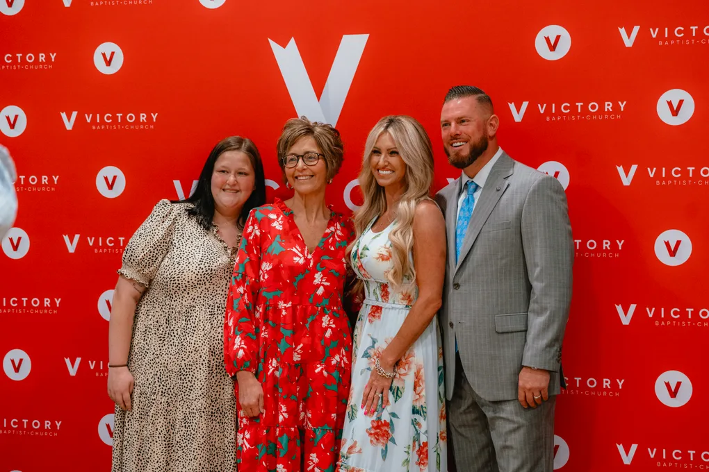 Pastor CT and Becky with two guests at the red Victory backdrop in the hospitality room