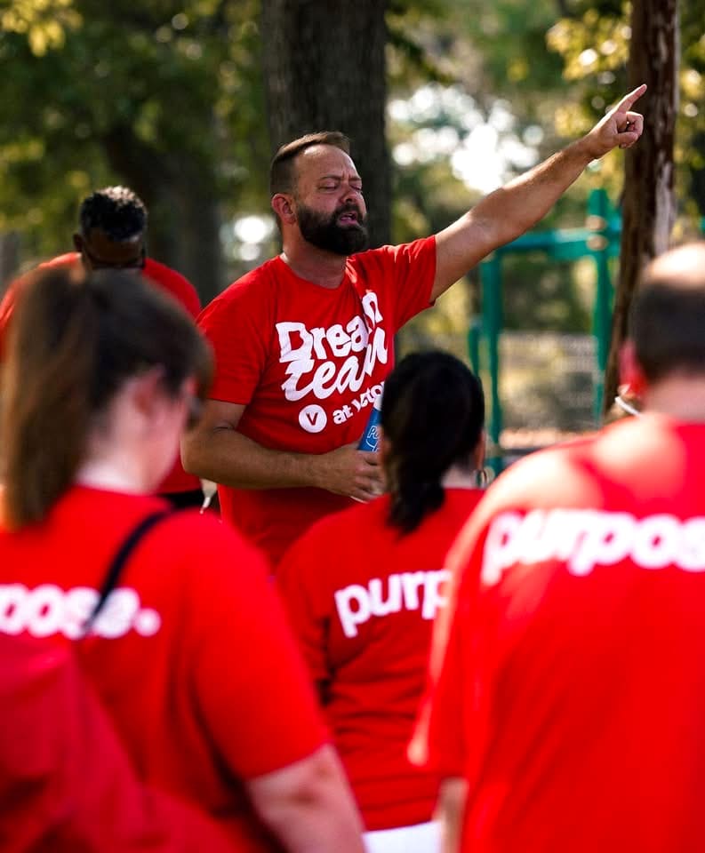 A Dream Team leader in a red Dream Team shirt pointing skyward as he speaks to the team in their purpose shirts at an outdoor serve day