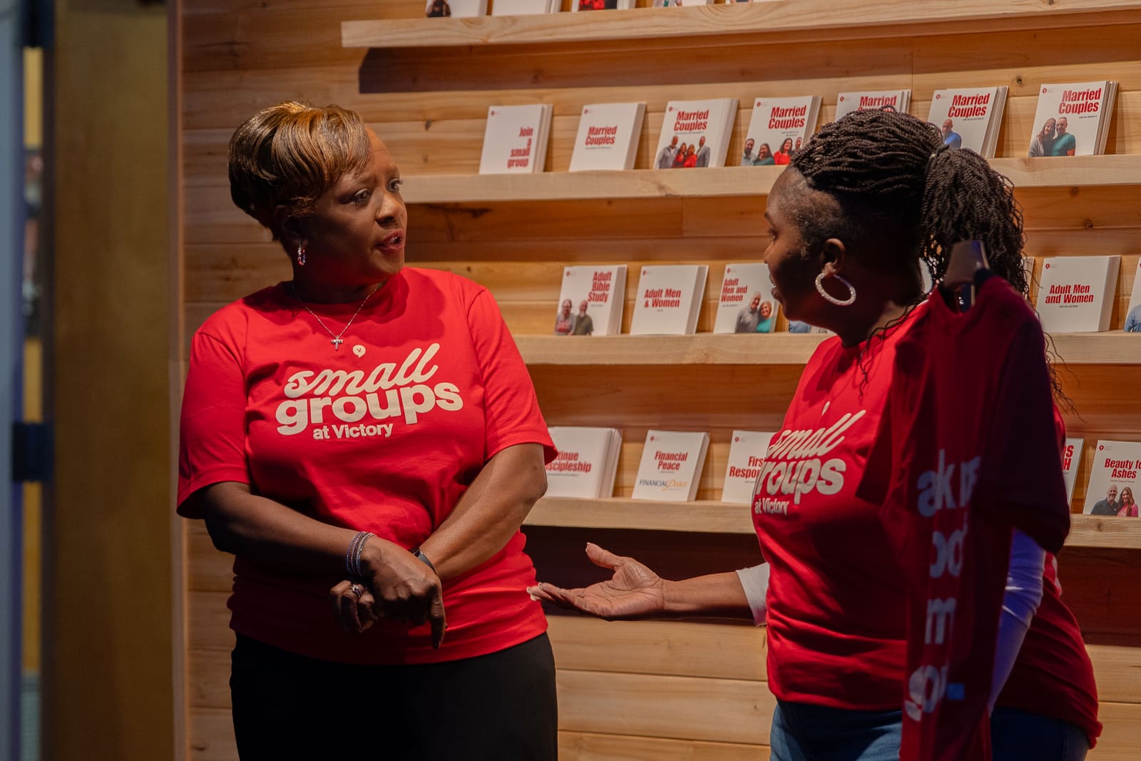 Two Small Groups team members in red Victory shirts talking at the curriculum wall