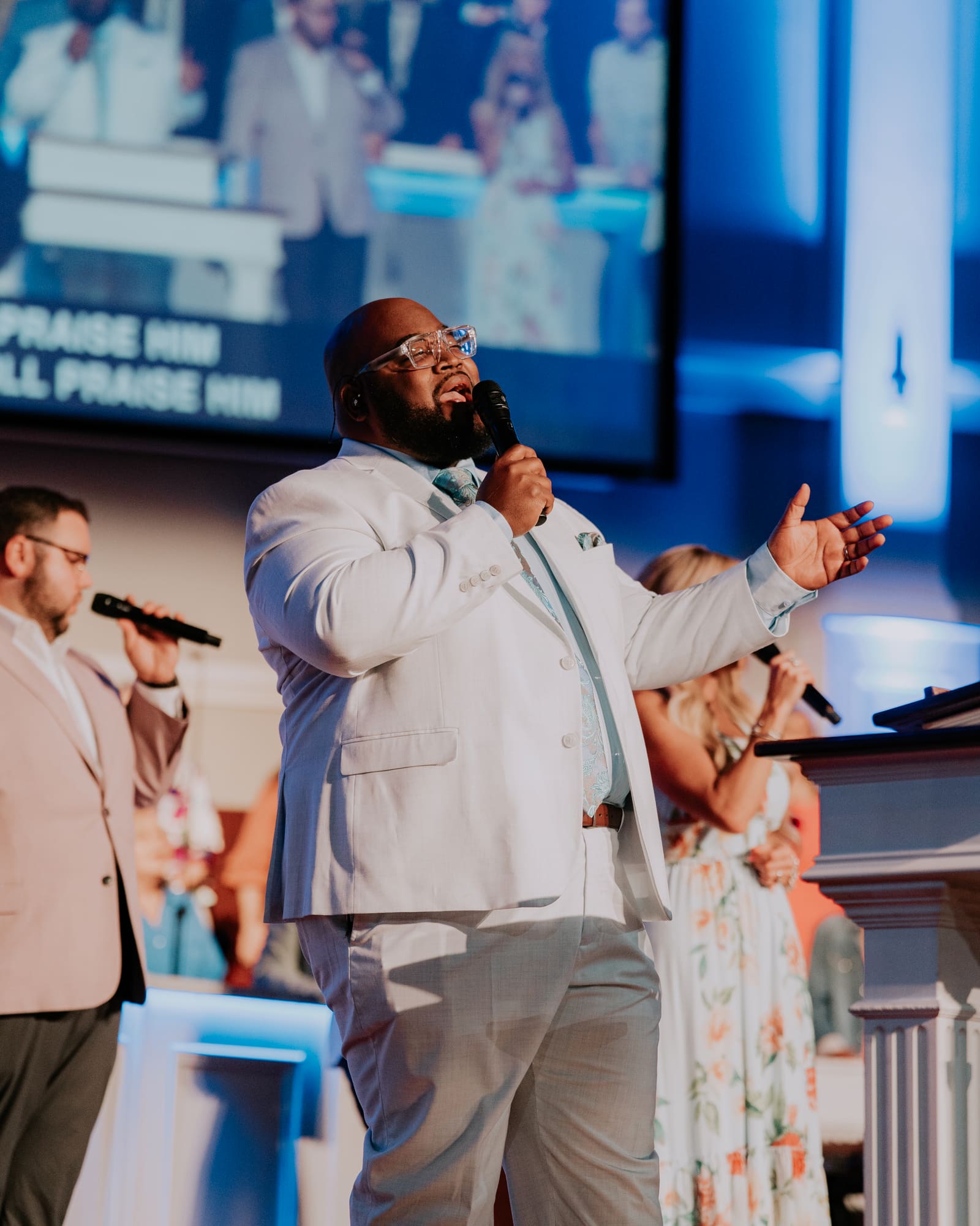 A worship leader in a light suit singing into a handheld mic from the platform, his arm stretched wide, the worship team behind him